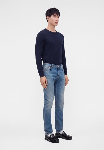 Jeune homme debout portant une chemise bleu marine à manches longues, un jean bleu clair, des chaussettes blanches et des mocassins noirs sur un fond blanc uni.