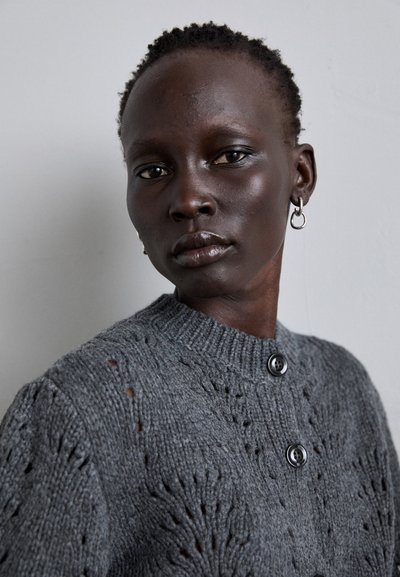 Grauer gestrickter Pullover mit einem strukturierten Muster, rundem Ausschnitt und zwei schwarzen Knöpfen auf der Vorderseite, getragen von einem Model mit kurzen Haaren.