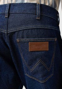 Jeans de mezclilla azul oscuro con un parche de cuero marrón que presenta "Wrangler" y costuras doradas en contraste; bolsillo trasero con un diseño distintivo.