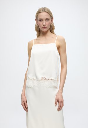 Femme blonde portant une robe blanche sans manches avec un galon en dentelle autour de la taille, debout devant un fond clair uni.