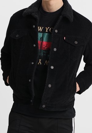Mann trägt eine schwarze Cordjacke mit Sherpa-Kragen über einem schwarzen T-Shirt mit New York-Design.
