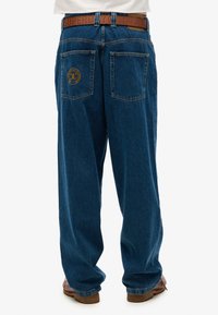 Jeans de mezclilla azul con un corte relajado, que cuentan con dos bolsillos traseros y un parche de logo en el bolsillo derecho. Completos con un cinturón marrón.