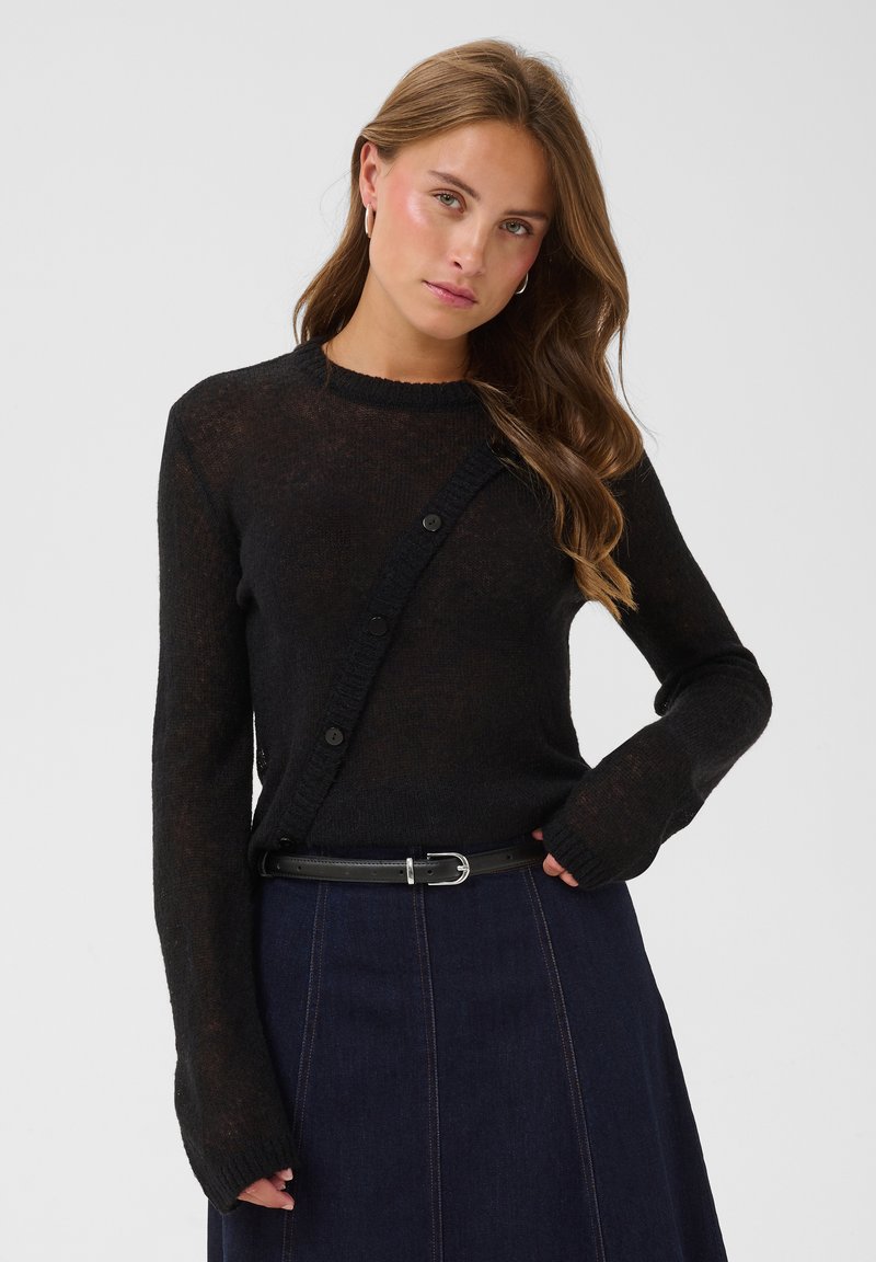 Jeune femme aux longs cheveux bruns porte un pull noir boutonné, une ceinture sombre et une jupe bleu marine, posant avec une main sur la hanche.