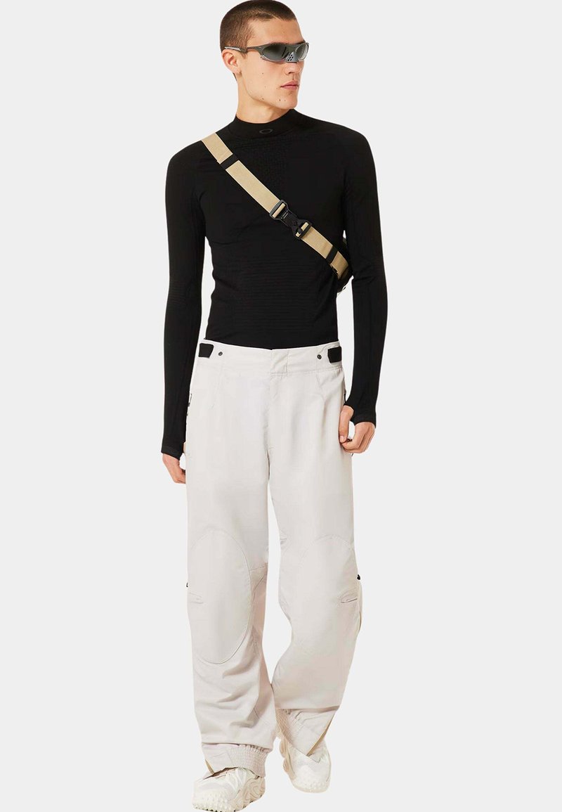 Schwarzes Langarmoberteil aus strukturiertem Stoff, weiße lockere Cargo-Hose mit runden Knie-Patches und beigem Umhängegurt. Graue Sonnenbrille.