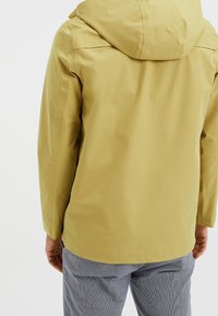 Yellow waterproof jacket with a hood, featuring a smooth texture and minimalistic design. Worn over gray patterned pants.