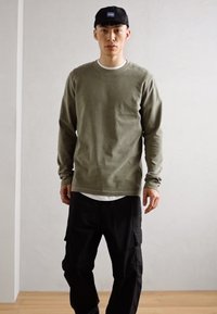 Camisa de manga longa oliva, com gola redonda, mistura de algodão, combinada com calças cargo pretas, textura suave, ajuste casual, usada com um boné escuro.