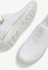Baskets blanches sans lacets en tissu maille texturée, semelles épaisses en caoutchouc avec une forte empreinte zigzag et un passant à l'avant supérieur.