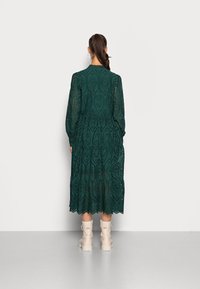 Lång klänning i smaragdgrönt med intrikata utskärningsmönster, långa ärmar, ett lager av volanger på fållen och en figursydd midja, tillsammans med beige ankelstövlar.