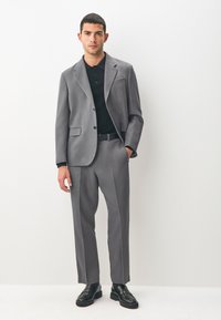 Costume gris sur mesure avec une veste structurée et un pantalon à devant plat. Associé à un polo noir et des chaussures en cuir noir. Texture de tissu lisse.