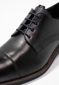 Chaussure habillée en cuir noir avec une finition lisse, dotée de six œillets, d'un accent texturé sur le talon et de coutures de semelle bleues contrastantes.