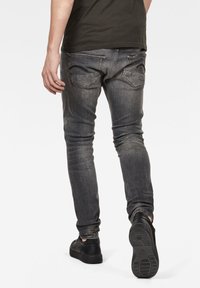 Vaqueros de mezclilla gris con un corte ajustado, que presentan un sutil desgaste, bolsillos traseros con acentos cosidos y un acabado usado. Se combinan con zapatillas negras.