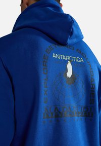 Homme portant un sweat à capuche bleu avec un dessin de pingouin et un globe, ainsi que le texte "Explore Beyond Boundaries Antarctica 1987 2023 Napapijri Geographic" au dos.