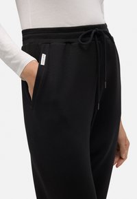 Pantalones de chándal negros con cintura ajustable, bolsillos laterales y una pequeña etiqueta de logo en la cadera. Tejido suave con un corte holgado.