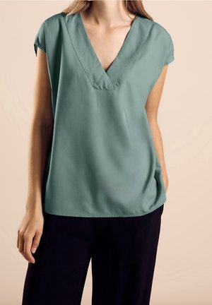 Vrouw die een loszittende groene V-hals blouse met korte mouwen en zwarte broek draagt, staand tegen een beige achtergrond.