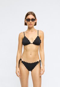 Μαύρο μπικίνι με διακοσμητικές σ scalloped; κορυφή τριγώνου και κάτω μέρος με πλευρικά κορδόνια. Το υλικό φαίνεται λείο και ελαστικό. Συμπεριλαμβάνει λεπτές ιμάντες και ελάχιστο μεταλλικό εξάρτημα.