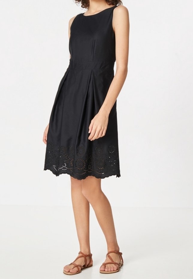 Femme portant une robe noire sans manches avec un ourlet floral découpé au laser et des sandales plates marron à brides, se tenant devant un fond blanc uni.
