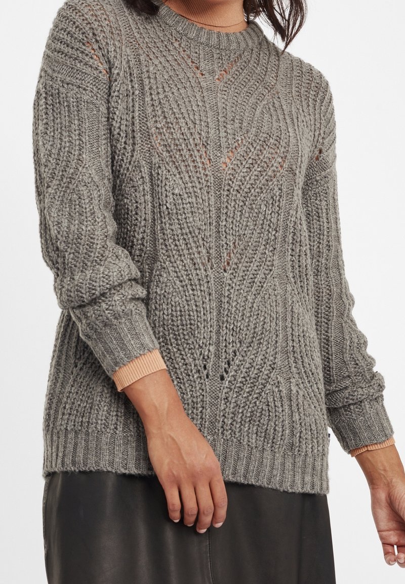 Gros plan d'une personne portant un pull en maille épaisse gris avec des motifs complexes par-dessus un haut à manches longues pêche et une jupe noire.