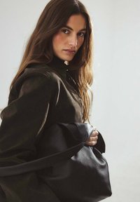 Sort læderhåndtaske med blød tekstur og en bred skulderrem, holdt tæt. Modellen bærer en mørkegrøn jakke. Lys baggrund.