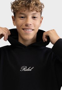 Juodas džemperis su ribota tekstūra, ant krūtinės išsiuvinėtu baltu "Rebel" logotipu. Kapota šiek tiek užtrauktas į priekį.