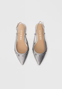 Zapatos planos tipo slingback en color plateado con punta puntiaguda, adornados con tachuelas decorativas y plantillas beige con la etiqueta "GUESS", exhibidos sobre un fondo blanco.