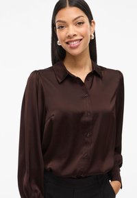 Smeđa satenasta košulja dugih rukava s ovratnikom, glatke teksture. Ima suptilne nabore na ramenima i dugmad u odgovarajućoj boji.