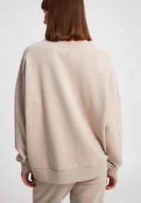 Sweatshirt beige con vestibilità ampia, collo a coste e maniche lunghe con polsini. Presenta un piccolo logo ricamato sul retro. Texture del tessuto morbida.