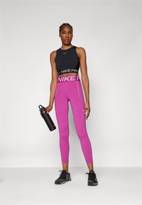 Juodas sportinis kūno prigludęs topas su "NIKE PRO" logotipu, derinamas su ryškiai rožiniais tamprėmis su prekės ženklo juostele. Modelis laiko juodą vandens butelį.