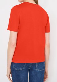 Personne portant une chemise rouge unie à manches courtes et un jean bleu, vue de dos sur un fond blanc.