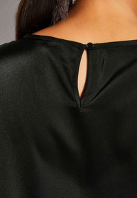 Schwarzes Satin-Oberteil mit einer Schlüsselloch-Detail am Rücken, das mit einem runden Knopf gesichert ist. Der Stoff hat eine glatte, glänzende Textur.