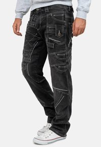 Pantaloni cargo in denim nero con numerose tasche, cuciture bianche a contrasto e vestibilità rilassata. Presentano dettagli in metallo decorativi.