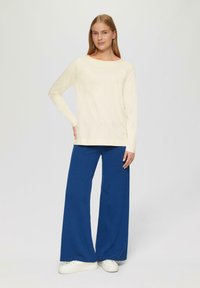 Crème lange mouwen top in combinatie met een wijd uitlopende blauwe broek. De textuur lijkt zacht, met een relaxte stijl en een eenvoudige snit. Witte sneakers maken de look af.