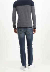 Man med en marinblå och grå randig långärmad skjorta, urblekta blå jeans och vita sneakers som står vänd bort mot en vit bakgrund.