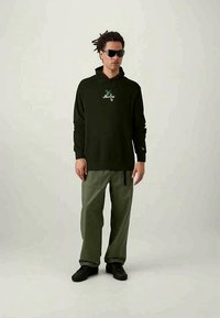 Tumši zaļš kapucis ar priekšējo logotipu, kombinēts ar gaiši zaļām kravas biksēm, kas ir ar sānu kabatu, valkāts ar melnām kurpēm un saulesbrillēm.