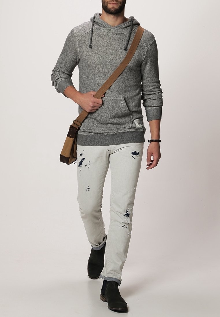Homme marchant portant un sweat à capuche gris, un jean clair usé, des bottes noires et un sac bandoulière marron avec une large sangle.