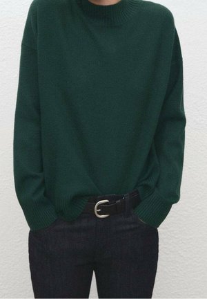 Personne portant un pull en maille vert foncé rentré dans un pantalon foncé avec une ceinture noire, les mains dans les poches, devant un mur clair uni.