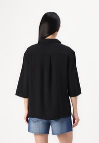 Black collared shirt with three-quarter sleeves and a relaxed fit, featuring a subtle back pleat, paired with denim shorts.