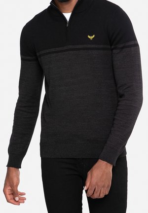 Mężczyzna ubrany w ciemnoszary i czarny sweter zapinany na zamek do ćwiartki z małym żółtym emblematem na lewej piersi, w połączeniu z czarnymi spodniami.