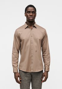 Πουκάμισο με μακριά μανίκια σε ανοιχτό καφέ, λείο ύφασμα με γιακά κουμπωτό και διακριτική κεντημένη στάμπα στο στήθος.