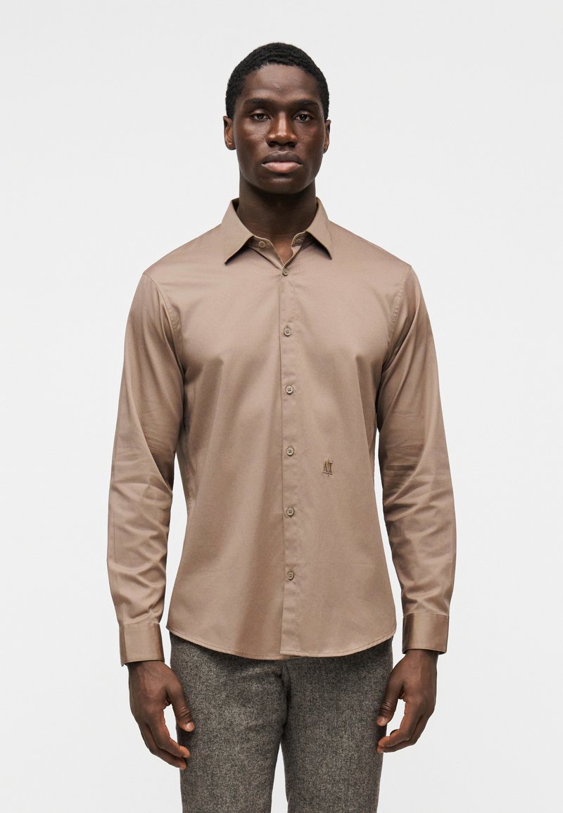 Πουκάμισο με μακριά μανίκια σε ανοιχτό καφέ, λείο ύφασμα με γιακά κουμπωτό και διακριτική κεντημένη στάμπα στο στήθος.