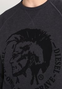 Mörkgrå sweatshirt med en stor svart grafik av en Mohawk-frisyr i profil, med texten "DIESEL" runt om. Tillverkad av mjukt material.