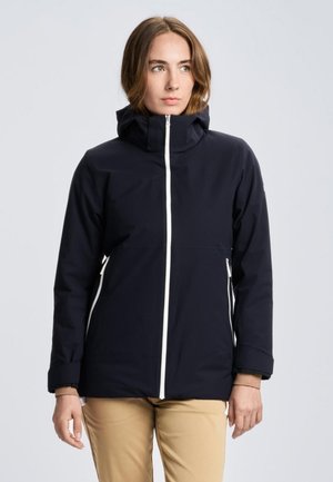 Modelo con una chaqueta azul marino oscuro de cuello alto, cremallera blanca delante y cremalleras en los bolsillos, combinada con pantalones beis sobre un fondo liso.