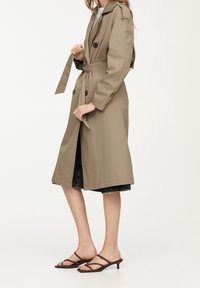 Femme ajustant la ceinture d'un trench-coat beige longueur genou, portant une jupe en denim noir et des sandales à talons brunes avec lanières.