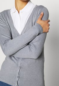 Cardigan gris de punto con diseño de escote en V, cierre de botones en la parte delantera y mangas largas, superpuesto sobre una camisa blanca con cuello.