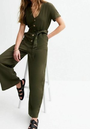 Kvinde iført olivengrøn jumpsuit med knapper, korte ærmer, bælte i taljen, vide ben og sorte sandaler med stropper, siddende på en hvid skammel.