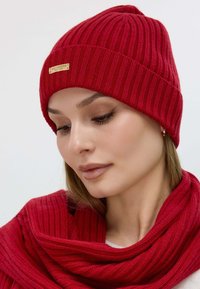 Red ribbed knit beanie with a small gold logo tag, paired with a matching red ribbed knit scarf. Both have a soft, textured appearance.