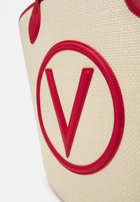 Vävd halminköpsväska med en krämfärgad bakgrund, röda läderdetaljer och en framträdande cirkulär logotyp med en stiliserad "V" i mitten.