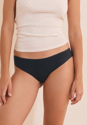 Vrouw draagt zwarte bikini-ondergoed en een lichtbeige mouwloos topje tegen een neutrale achtergrond.