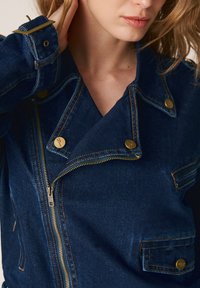 Kobieta ubrana w ciemnoniebieską kurtkę dżinsową z złotymi guzikami i zamkiem, ręka dotyka szyi, widoczne włosy do ramion w kolorze blond.