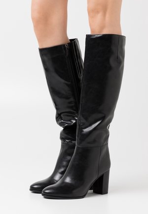 Bottes noires montantes en cuir brillant avec un talon bloc et une fermeture éclair latérale. Le design présente une partie supérieure froncée pour une texture supplémentaire.