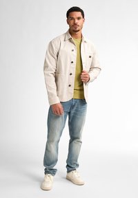 Light beige canvas jacket with button closure, two front pockets, layered over a green top, paired with light blue jeans and cream sneakers.